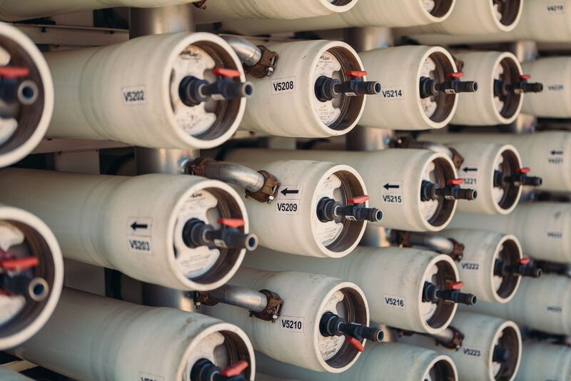 A close-up view of multiple rows of industrial reverse osmosis membrane filters. The cylindrical white vessels are arranged in parallel rows. The vessels have black and red connectors or ports on their ends, and small directional arrows are visible on their surfaces. The image shows the systematic arrangement of these water treatment components in what appears to be a water purification facility.